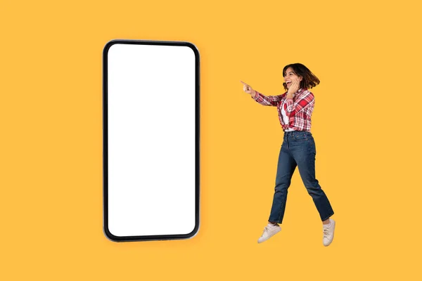 感情的にかなり中東の若いです女性でカジュアルなジャンプとポーティングで巨大な携帯電話とともにホワイトブランクスクリーン上の黄色のスタジオの背景 ジェスチャー 素晴らしいモバイルアプリ モックアップ — ストック写真