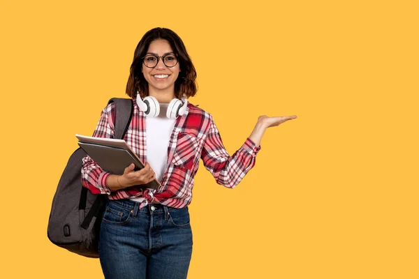黄色のスタジオの背景にコピースペースを示すカジュアルな学生で積極的な魅力的な若いアラブ女性は 眼鏡をかけて バックパックを運ぶ ノートパソコンや本を保持 — ストック写真
