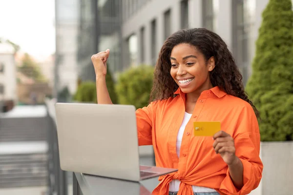 Happy young black female with credit card looks at computer, raises hand up, making gesture of victory and success in city. People emotions with gadget, social distancing, business and online shopping