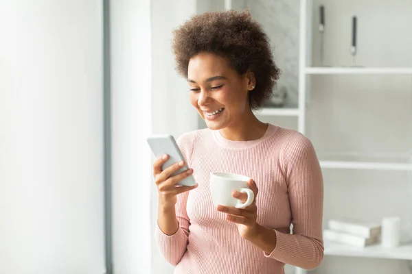 快乐的千年非洲裔美国女人在电话里聊天 在客厅里喝一杯最喜欢的饮料 申请博客和社交网络 在家中免费上网 — 图库照片