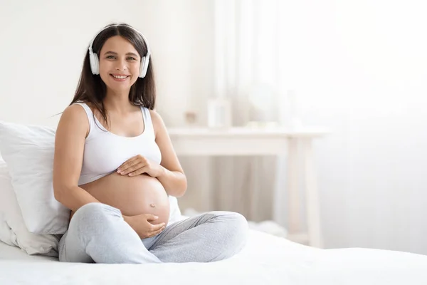 幸せな笑顔の若い妊婦リラックスし 音楽を聴きながら自宅でベッドに座って 彼女の大きなおなか 白い寝室のインテリア コピースペースのパノラマに触れる 子宮の概念の赤ん坊のための音楽 — ストック写真
