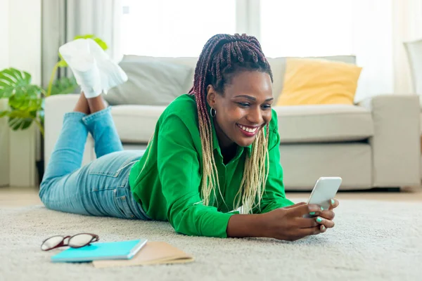 Excited black woman lying on floor with smartphone, working remotely or video chatting at home, free space. Positive african american lady watching movie, having online meeting