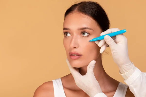 Plastic surgeon drawing lines on young womans face for cosmetic operation on beige studio background, copy space. Beautiful lady getting ready for facelift