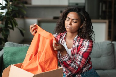 Gayri resmi bakışlar içinde mutsuz, genç siyahi bir kadın. Kıyafetlere bakıyor, paketleri oturma odasının iç tarafına açıyor. Çevrimiçi alışveriş, hata teslimatı, kötü satın alma kalitesi vd-19