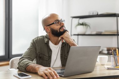 Düşünceli Latin erkek serbest çalışan ev bilgisayarında çalışırken bir şey düşünüyor, masada oturuyor ve mutlu bir yüz ifadesiyle başka yere bakıyor, boşluğu kopyalıyor.