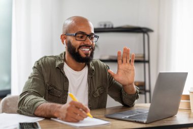 Telekonferans ve telekomünikasyon. Masada oturan mutlu latin adam dizüstü bilgisayarında video görüşmesi yapıyor, web kamerasına el sallıyor. Sanal toplantıda kulaklık takan olumlu bir erkek.