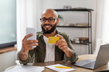 Evden online alışveriş yaparken kredi kartı ve akıllı telefon kullanan mutlu bir zenci, dizüstü bilgisayarla masada oturuyor. Adam telefonda banka uygulaması üzerinden ödeme yapıyor. E-ticaret kavramı