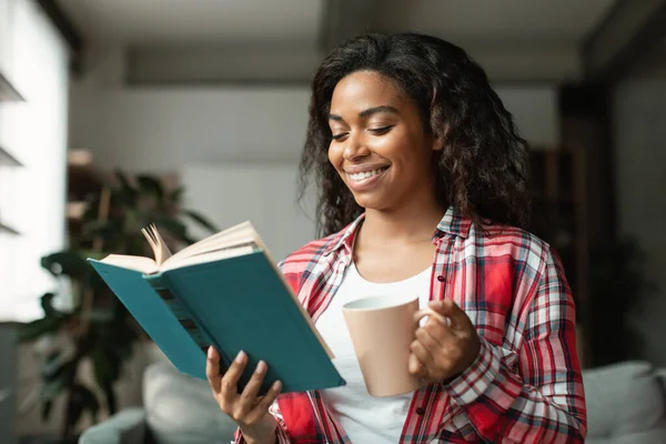 Cheerful smart pretty black young woman student reading book, enjoy cup of hot favorite drink, study and spare time in living room interior. Literature, hobby, coffee break at home alone, covid-19