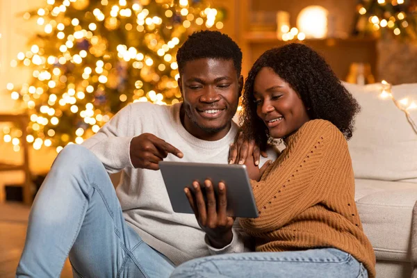 若いアフリカ系アメリカ人男性の笑顔女性にタブレットを表示 ビデオ通話を持っています クリスマスツリーとガーランドとリビングルームのインテリアで休日をお楽しみください 新年のリモートのお祝い おめでとうのためのアプリ — ストック写真