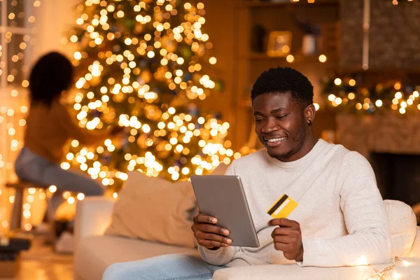 タブレットやクレジットカードを持つ幸せな千年紀の黒人男性は オンラインショッピングを楽しみ 妻はガーランドと部屋のインテリアでクリスマスツリーを飾ります 新年への準備 購入注文 アプリと休日の販売 — ストック写真