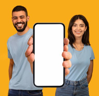 Harika bir uygulama. Beyaz Ekran ile Büyük Boş Akıllı Telefon Gösteren Mutlu Arap Çifti, Orta Doğulu Gülümseyen Adam ve Kadın, Sarı Arkaplan 'ın üzerinde dururken Ellerinde Boş Cep Telefonu Tutuyor,