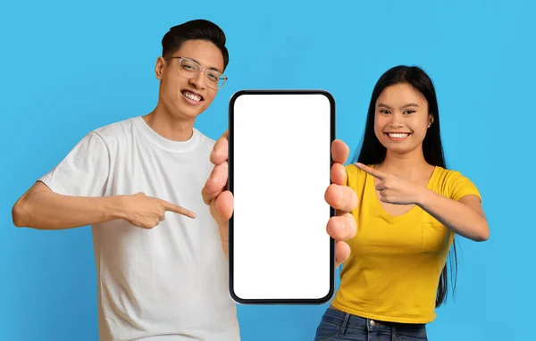 不错的提议 年轻的亚洲夫妇手里拿着并指着大大的空白智能手机 快乐的男人和女人拿着空白的手机进行模仿 站在蓝色的背景上 拼凑在一起 — 图库照片