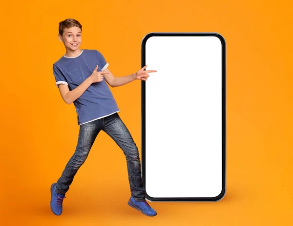 伟大的应用 快乐的男孩儿带着空白白屏在大智能手机前指点 积极的青春期男孩演示移动应用程序设计的复制空间 站在橙色背景上 — 图库照片