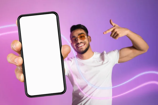 陽気な千年紀のアラブ人の男でサングラスと白のTシャツを指している現代の携帯電話で白い空の画面 親指を表示 推奨しています素敵なモバイルアプリ モックアップ コラージュ — ストック写真