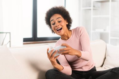 Mutlu bin yıllık Afro-Amerikan kadın telefonda online oyun oynayan, oturma odasının iç kısmında kanepede oturan. Kadın video izler, evde tek başına eğlenmek için uygulama kullanır, insanların duyguları...