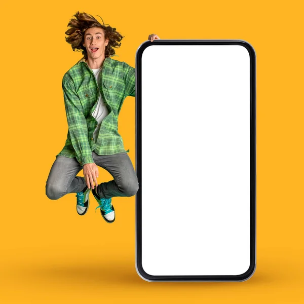 Amazing Offer. Excited Hipster Guy Jumping Near Big Blank Smartphone Over Yellow Background In Studio, Cheerful Young Male Demonstrating Copy Space For Mobile App Or Website Design, Collage
