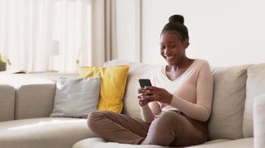 Çevrimiçi kazanma kavramı. Kapalı alanda çekilen Afro-Amerikan kadın akıllı telefondan kazanan mesajı okuyor, başarıyı kutluyor, evdeki kanepede oturuyor, yavaş çekim, takip çekimi, boş alan.