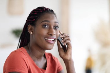 Evde cep telefonuyla konuşan güzel, gülümseyen siyahi bir kadının portresi. Neşeli, Afro-Amerikan bir kadının cep telefonu sohbetinden hoşlandığı yakın çekim.