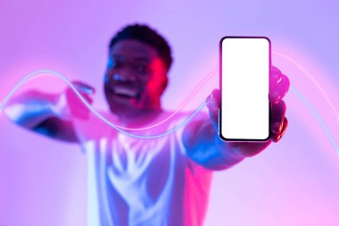 Beyaz ekranlı en yeni akıllı telefonu işaret eden, neon ışıkta poz veren, heyecan verici online teklif, taklit, kolaj öneren mutlu, genç ve çekici Afrikalı Amerikalı adam.