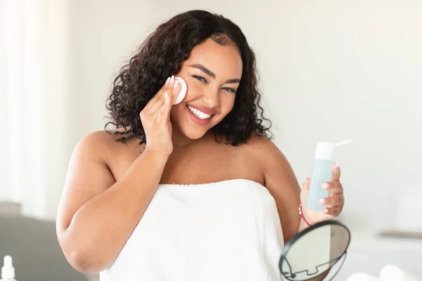 Attractive black body positive woman using facial toner and cotton pad for moisturising skin, happy lady making beauty treatments while sitting in bedroom at home, copy space