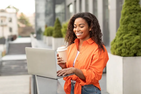 Smiling young black lady with cup of coffee to go chatting on computer, working remotely, has video call, meeting in city. Gadget and social distance for business and study outdoor and new normal