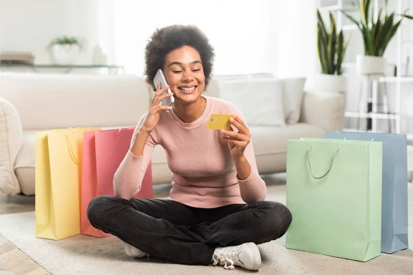 自宅でオンラインショッピング 購入の多くの袋を持つ幸せな千年のアフリカ系アメリカ人女性は クレジットカードを見て 電話で呼び出し 部屋のインテリアの床に座っています 良いニュース ゴシップ キャッシュバック — ストック写真