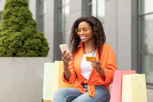 オンラインショッピングを楽しむクレジットカード パッケージ スマートフォンを備えた幸せなかなり若い黒人女性は 市内のベンチに座っています 大規模な販売と現代の金融アプリ キャッシュバックと顧客のための新しい通常 — ストック写真