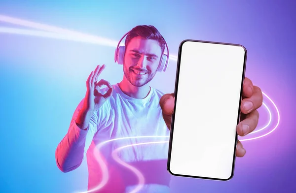 Przystojny Młody Człowiek Słuchawkami Bezprzewodowymi Pokazujący Telefon Komórkowy Białym Czystym — Zdjęcie stockowe
