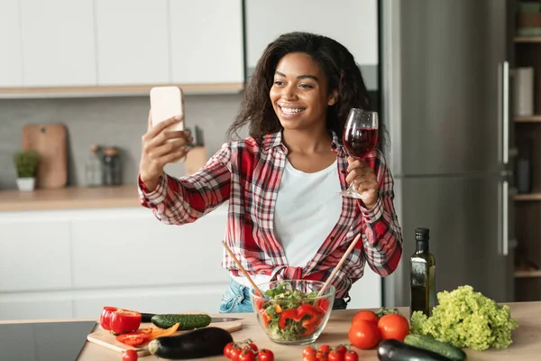 Smiling black pretty young woman in casual with salad and glass of wine makes selfie or has video call in modern kitchen interior with organic vegetable. Meeting remotely, app for food blog at home