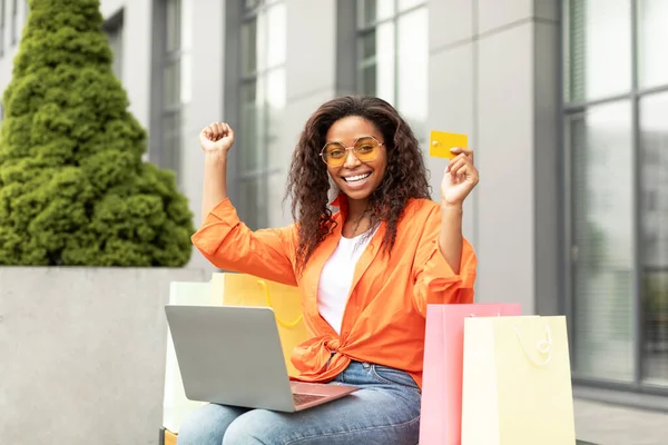 クレジットカード バッグ Pcを備えた満足の若い黒人女性は オンラインショッピングを楽しみ 手を上げ 素晴らしいオファー キャッシュバックで喜び 市内のベンチに座っています リモートで注文 成功し — ストック写真