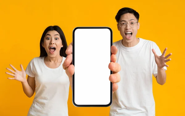 神奇的应用程序 令人惊讶的年轻亚洲男人和女人拿着大大的空白智能手机 带着白色屏风 感情用事的夫妻在惊奇中张开嘴 推荐网上优惠或移动广告 — 图库照片