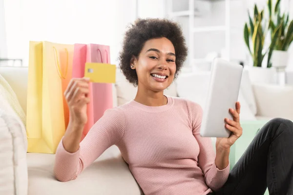 購入の多くのパッケージを持つ陽気な100万人のアフリカ系アメリカ人女性は リビングルームのインテリアにタブレットで座ってクレジットカードを示しています 自宅での販売 金融銀行 注文やオンラインショッピングのためのアプリ — ストック写真