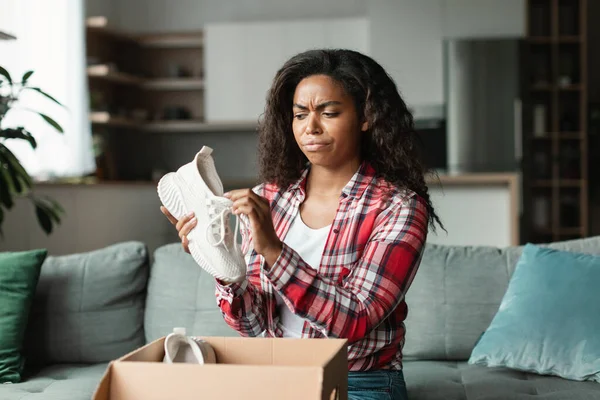 Sad displeased angry black young woman in casual unpacks parcel, looks at new sneakers in living room interior. Shopaholic emotions from online shopping, mistake or damaged purchase and delivery order
