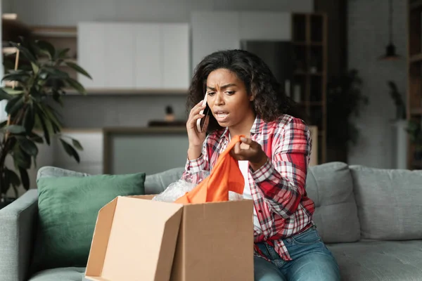 Angry black young woman in casual unpacks parcel, calls by smartphone, swears with store in living room interior. Problems with online shopping, mistake of delivery, purchase, wrong or damaged order