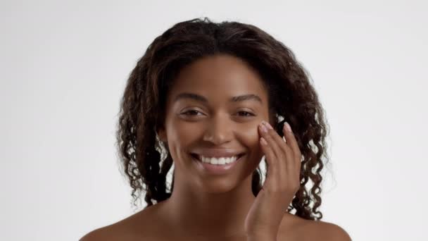 セルフケアの手順 若いです幸せなアフリカ系アメリカ人女性摩擦クリーム上の頬 彼女の頬を甘やかす カメラに笑みを浮かべて 白の背景 スローモーション — ストック動画
