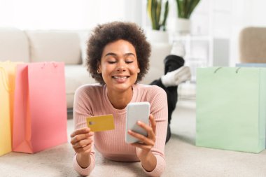 Bir sürü paketi olan gülümseyen Afro-Amerikalı kadın kredi kartı kullanıyor, akıllı telefon kullanıyor, oturma odasının içinde yerde yatıyor. Nakit para, çevrimiçi alışveriş, satış ve ev siparişi uygulaması