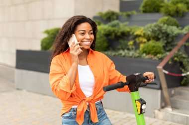 Elektrikli scooter kullanan neşeli siyah kadın akıllı telefon, konuşma, iletişim, şehir parkında yürümekten hoşlanıyor. Kentsel hizmet, dedikodu, iyi haber, cihazla seyahat ve modern çevre taşımacılığı