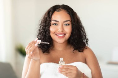 Cilt bakım kozmetiği. Güzel, siyah, iri yarı, elinde yüz serumu olan ve kameraya gülümseyen bir kadın. Evde güzellik tedavisinden zevk alıyor.