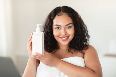 Güzellik reklamı. Mutlu siyah tombul kadın elinde nemlendirici vücut losyonu şişesiyle kameraya gülümsüyor, yeni kozmetik reklamlar yapıyor, havluya sarılı duruyor.