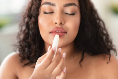 Genç Afro-Amerikan tombul bayan dudak kremi sürüyor, nemlendiriyor ve cilt bakımı yapıyor, yakın çekim. Güzel siyah kadın kozmetik ürünler kullanıyor, dudaklarına dikkat ediyor.