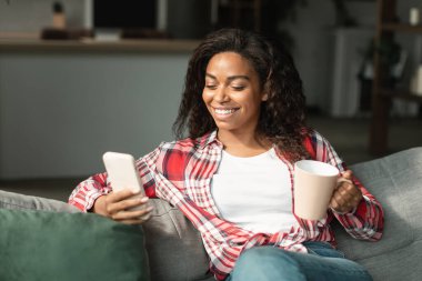 Neşeli siyahi bir kadın, bir bardak içkiyle telefonda sohbet ediyor, blog okuyor, minimalist oturma odasında rahat ediyor. Cihazda uzaktan iletişim, sosyal ağlar ve evde yeni uygulama