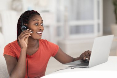 Telekomünikasyon konsepti. İçişleri Bakanlığı 'nda dizüstü bilgisayarda çalışan Afro-Amerikan kadın kulaklık takan, masada oturan ve müşterisiyle çevrimiçi iletişim kurmak için bilgisayar kullanan siyah bir kadın.
