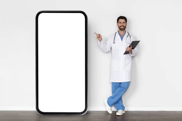 医疗服务 穿着制服的男医生站在医院白墙边的大空白智能手机屏幕前对着手指微笑 医生展示广告的复制空间 — 图库照片