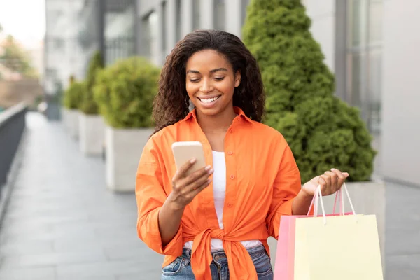 スマートフォンでカジュアルな入力で陽気千年黒の女性は 良いメッセージを読んで 市内での購入とパッケージを保持します オンラインショッピングデバイス 素晴らしい広告とオファー ライフスタイルと都市の散歩 — ストック写真