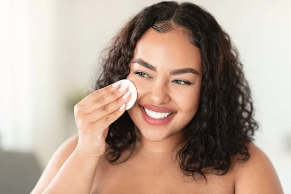 Closeup portrait of happy black oversize woman using cotton pads, erasing make up from her face, applying facial toner or cleansing milk, free space