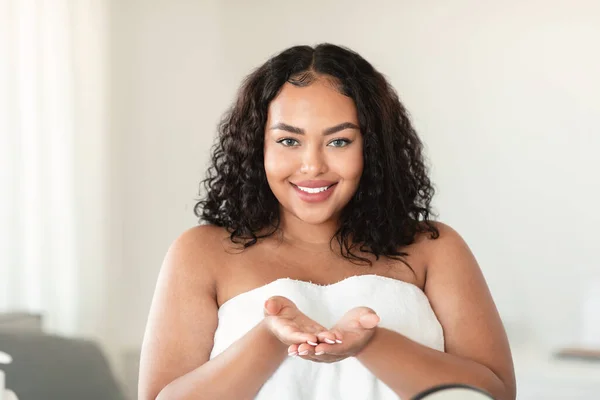 Attractive black chubby woman wrapped in bath towel demonstrating something on empty palms, showing invisible object and smiling at camera, free copy space