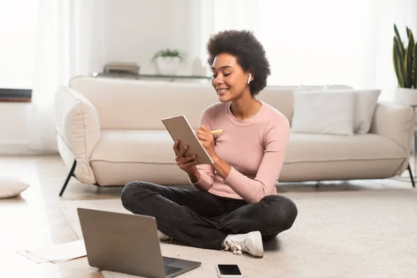 ワイヤレスヘッドフォンを備えた幸せな忙しい千年のアフリカ系アメリカ人女性はノートブックにメモを取り リビングルームのインテリアにラップトップ付きの床に座っています ビジネス ビデオレッスン 家庭での指導 — ストック写真