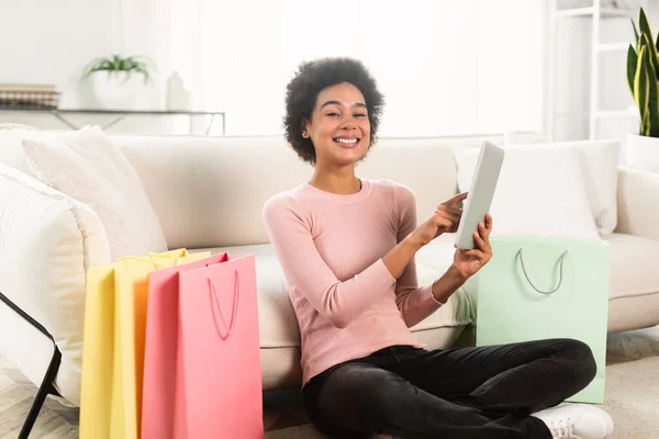 購入の多くの袋を持つ幸せな千年のアフリカ系アメリカ人の女性は 明るいリビングルームのインテリアでタブレットで床に座っています 巨大な販売のためのアプリ ファッションブログ キャッシュバック 注文やオンラインショッピング — ストック写真