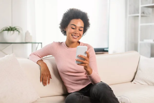 笑着漂亮的千年非洲裔美国女人在智能手机上聊天 坐在明亮客厅的沙发上 女士在家观看视频 阅读博客 上网和社交网络 — 图库照片
