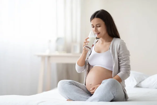 Long-haired young pregnant woman in comfy homewear sitting on bed, drinking milk at home, touching her big tummy, cozy white bedroom interior, copy space. Nutrition during pregnancy